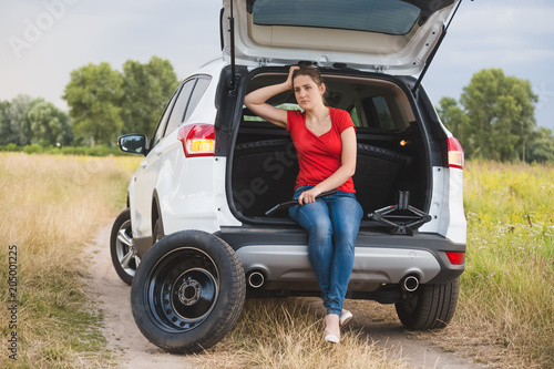 Young woman sitting in open car trunk and holding tools wor changing flat tire