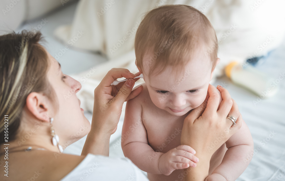 Young caring mother clearing baby son's ears from earwax with cotton swab Young caring mother clearing baby son's ears from earwax with cotton swab