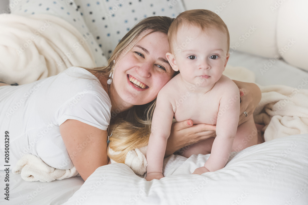 Toned portrait of laughing young mother lying on bed and embracing her baby son