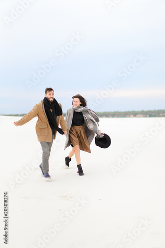 Smiling man and woman walking and holding hands in white monophonic background, wearing coats and scarfs. Concept of happy couple and seasonal photo session. photo