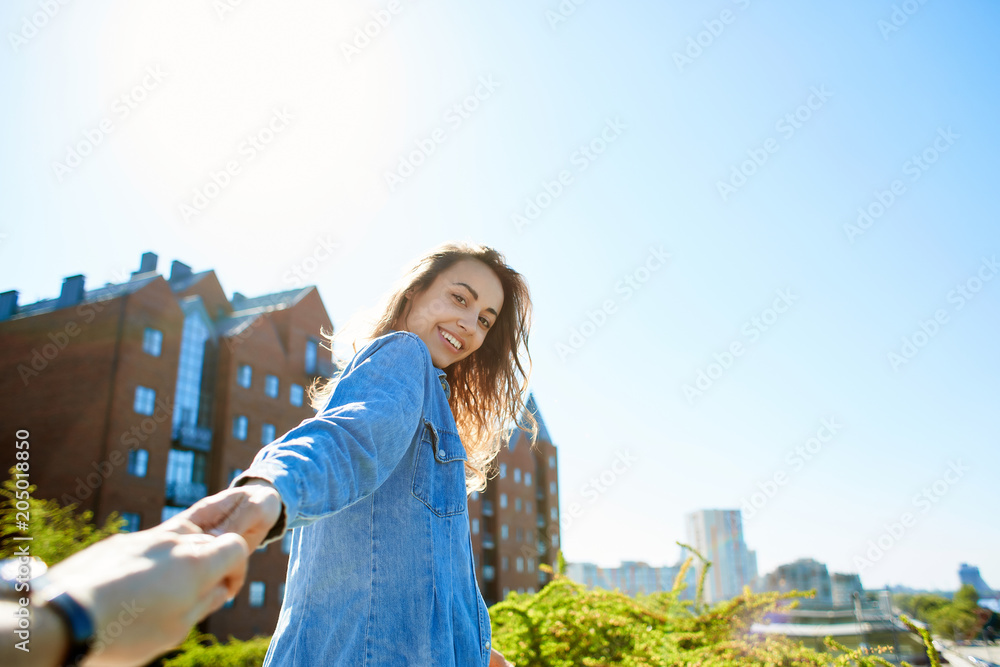 portrait of a young smiling attractive woman in jeans clothes at sunny day on the blue sky background. happy woman gives a hand to someone like follow me . first person view.