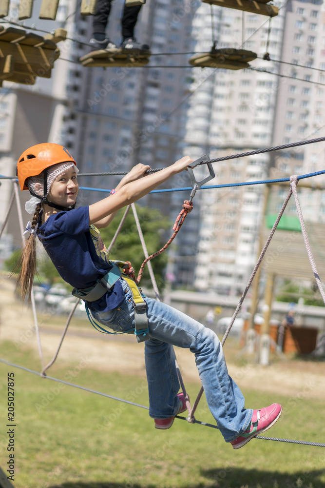 happy little girl is engaging in rock climbing in the cable car park. Little beautiful girl climbs on rope harness in summer city park. happy little girl is engaging in rock climbing in the cable car park. Little beautiful girl climbs on rope harness in summer city park.