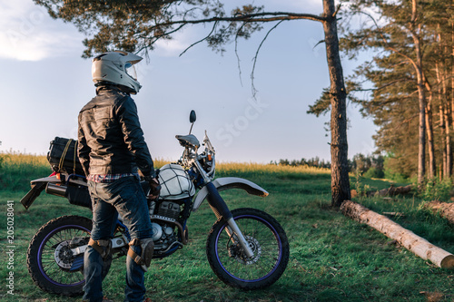 Fototapeta Naklejka Na Ścianę i Meble -  motorcyclist with his bike, sunset outdoor view, green grass and pine forest, off road motorcycle adventure, enduro, rider, dual sport road trip, travel concept