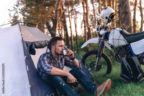 man is sitting in a tourist tent and holding metal cup in his hand, drink. Camping in forest, motorcycle touring, dual sport enduro, tent and off road adventure motorcycle, active life style concept