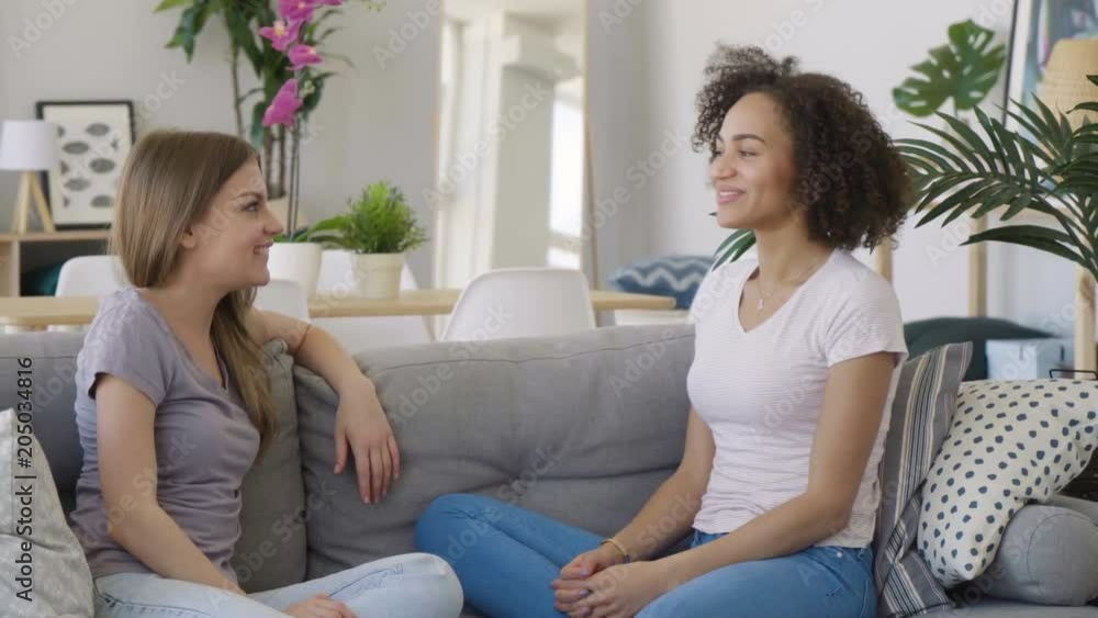 Two young attractive women caucasian blonde and brunette talk friendly and laugh cheerfully sitting on cozy coach in an airy spacious white room decorated in Scandinavian style in the morning Two young attractive women caucasian blonde and brunette talk friendly and laugh cheerfully sitting on cozy coach in an airy spacious white room decorated in Scandinavian style in the morning