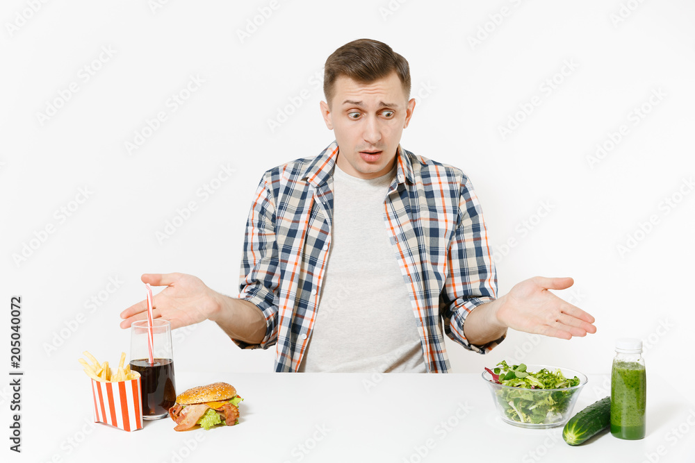 Man sits at table with green detox smoothies, salad in glass bowl, cucumber, burger, fries, cola in glass isolated on white background. Proper nutrition, healthy lifestyle, fast food, choice concept. Man sits at table with green detox smoothies, salad in glass bowl, cucumber, burger, fries, cola in glass isolated on white background. Proper nutrition, healthy lifestyle, fast food, choice concept.