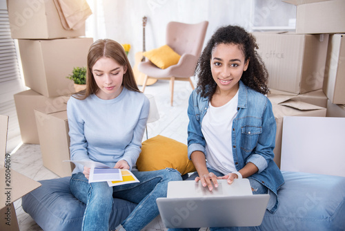 Convenient shopping. Charming female students sitting on the floor among boxes with their belongings and buying paint for new flat online photo