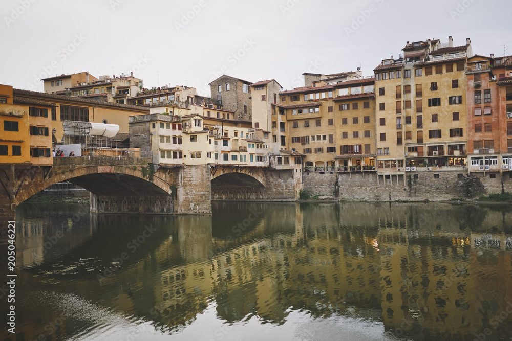 Florence, Italy. View of the Golden bridge and old houses on the waterfront of the river Arno. Cloudy sky. Houses are reflected in the water.