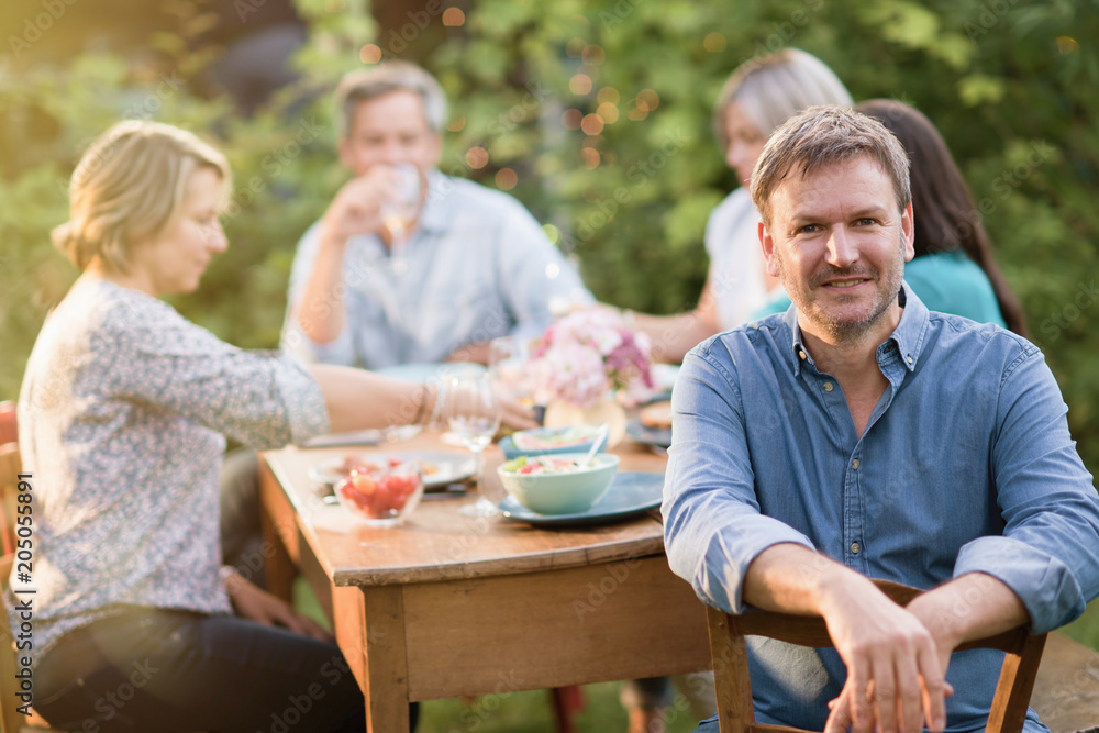 Looking at the camera, a man in his forties with a glass of wine in his hand sits at a table in the garden with his friends for dinner. Looking at the camera, a man in his forties with a glass of wine in his hand sits at a table in the garden with his friends for dinner.