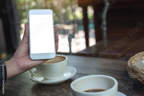 Mockup image of a hand holding and showing a white mobile phone with blank desktop screen with coffee cups on wooden table in cafe