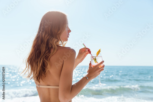 View from back on woman with long hair, holding a wine glass with cocktail, spending vacation on the beach, near the sea. photo