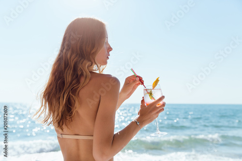 Canvas Print View from back on woman with long hair, holding a wine glass with cocktail, spending vacation on the beach, near the sea