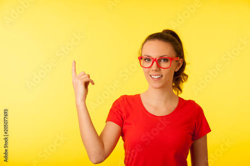 Young geek woman in red t shirt point in copy space over vibrant yellow background