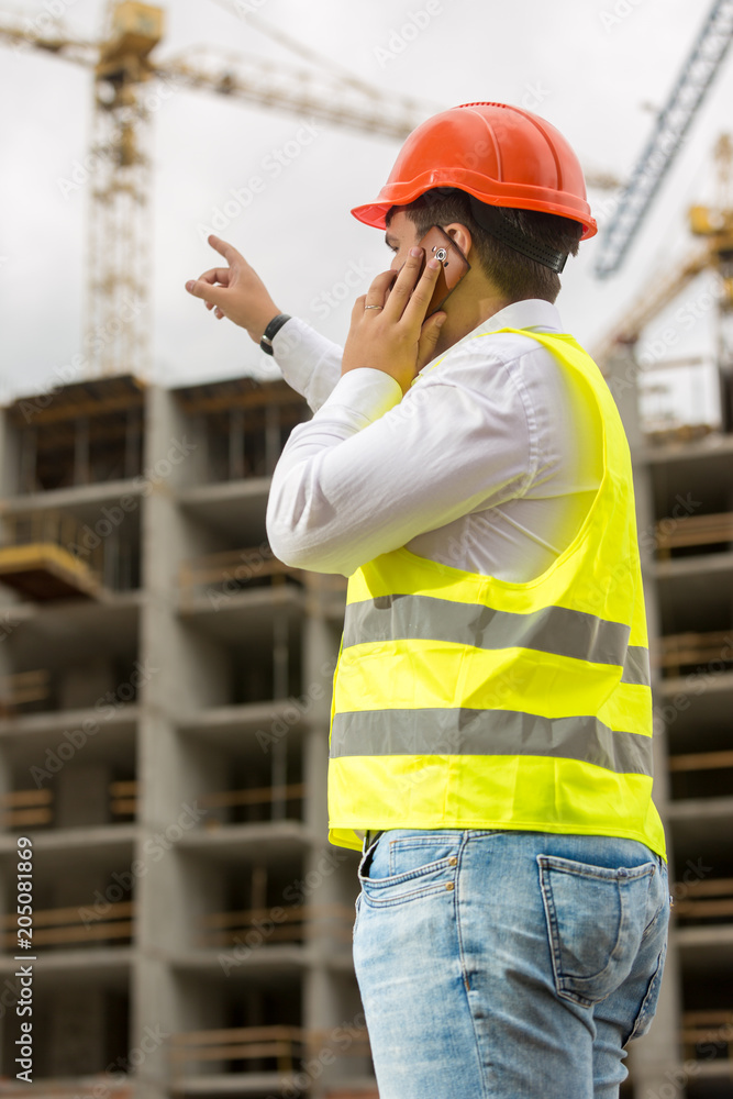 Young construction engineer in hardhat talking by phone and pointing at building under construction Young construction engineer in hardhat talking by phone and pointing at building under construction