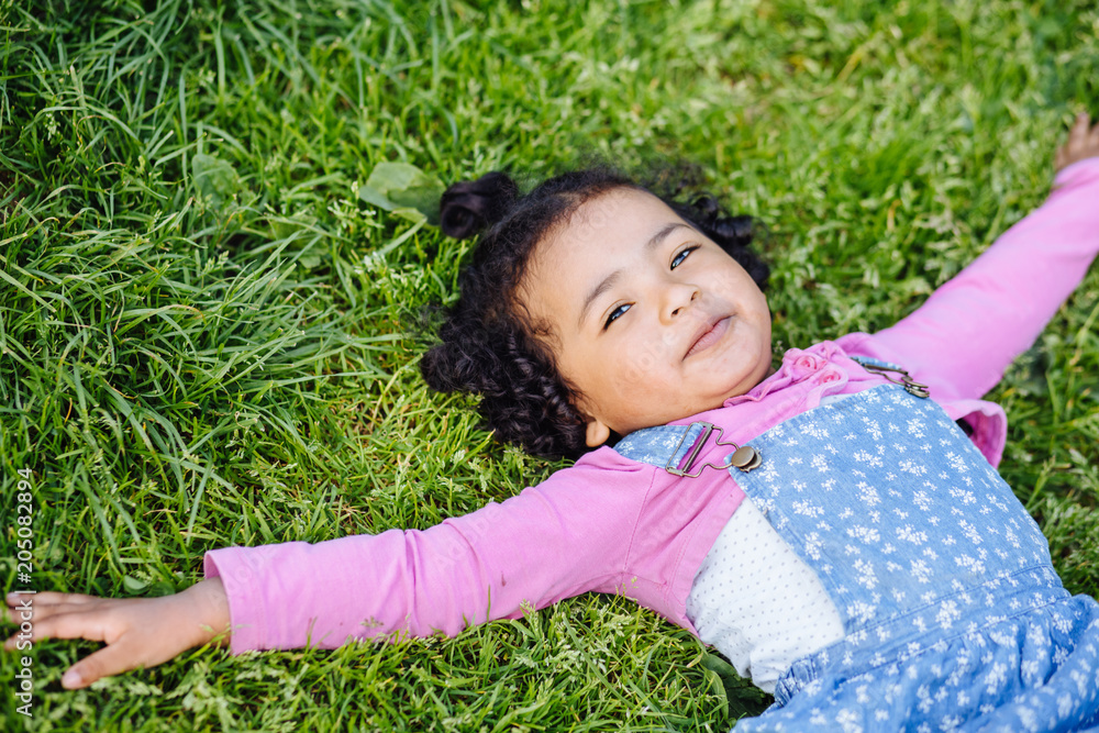 Portrait of dark skinned active hispanic or mexican toddler girl relaxing on green grass. Summer time weekend mood. Caucasian child dressed in casual clothing: blue denim sarafan, pink shirt. Portrait of dark skinned active hispanic or mexican toddler girl relaxing on green grass. Summer time weekend mood. Caucasian child dressed in casual clothing: blue denim sarafan, pink shirt.
