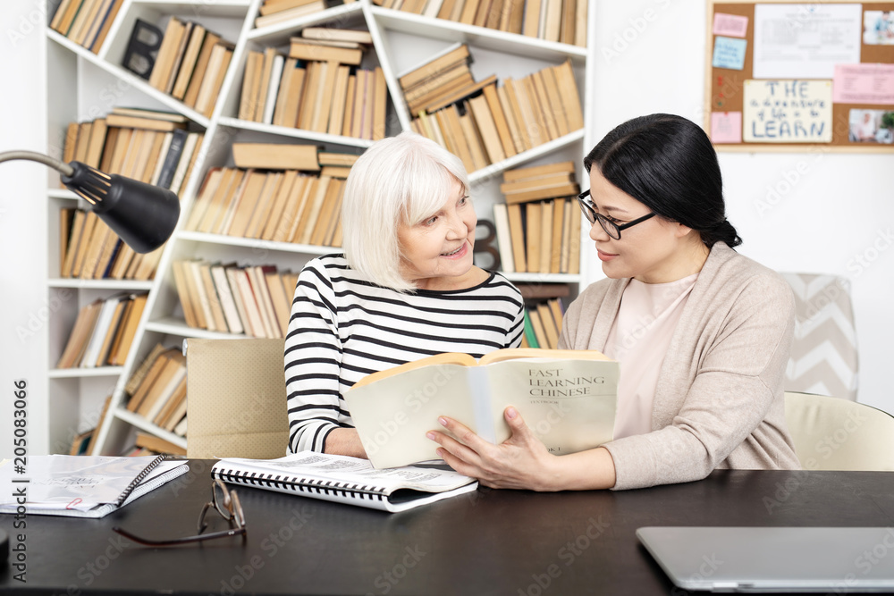 Full explanation. Appealing tutor carrying book while senior woman asking question Full explanation. Appealing tutor carrying book while senior woman asking question