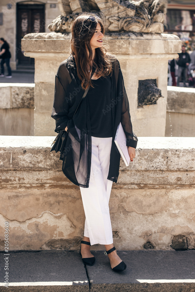beautiful stylish woman standing in sunny city street, holding magazine. happy hipster girl in fashionable outfit, relaxing and enjoying day in european city . beautiful stylish woman standing in sunny city street, holding magazine. happy hipster girl in fashionable outfit, relaxing and enjoying day in european city .
