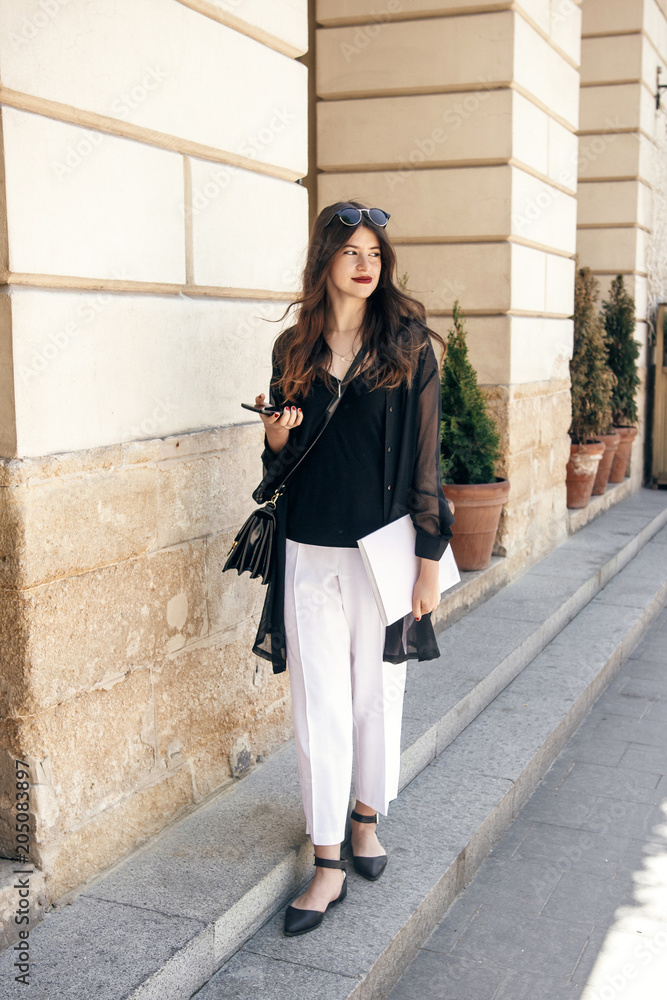 beautiful stylish woman walking in sunny city street, holding phone and magazine. happy hipster girl dressed in fashionable outfit, posing in european city beautiful stylish woman walking in sunny city street, holding phone and magazine. happy hipster girl dressed in fashionable outfit, posing in european city
