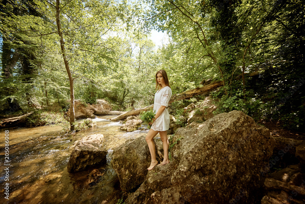 Young pretty woman in a white dress walking and posing in a forest near the river