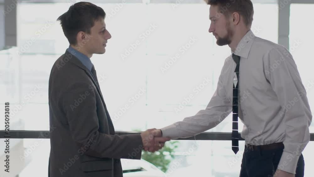 Two young businessmen is meeting, shaking hands in modern office, friendly professionals greeting, talking, walking along corridor of company. Concept: cooperation, entrepreneurship, conversation Two young businessmen is meeting, shaking hands in modern office, friendly professionals greeting, talking, walking along corridor of company. Concept: cooperation, entrepreneurship, conversation