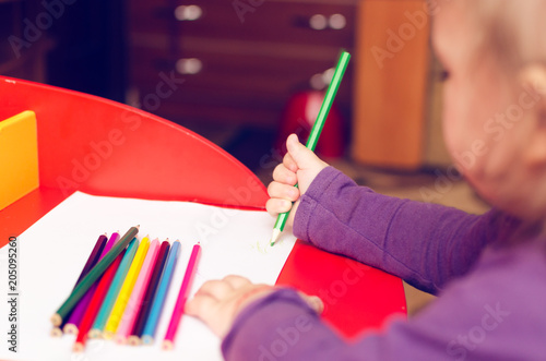 On the red table, children's hands with colored pencils and a blank sheet of paper
