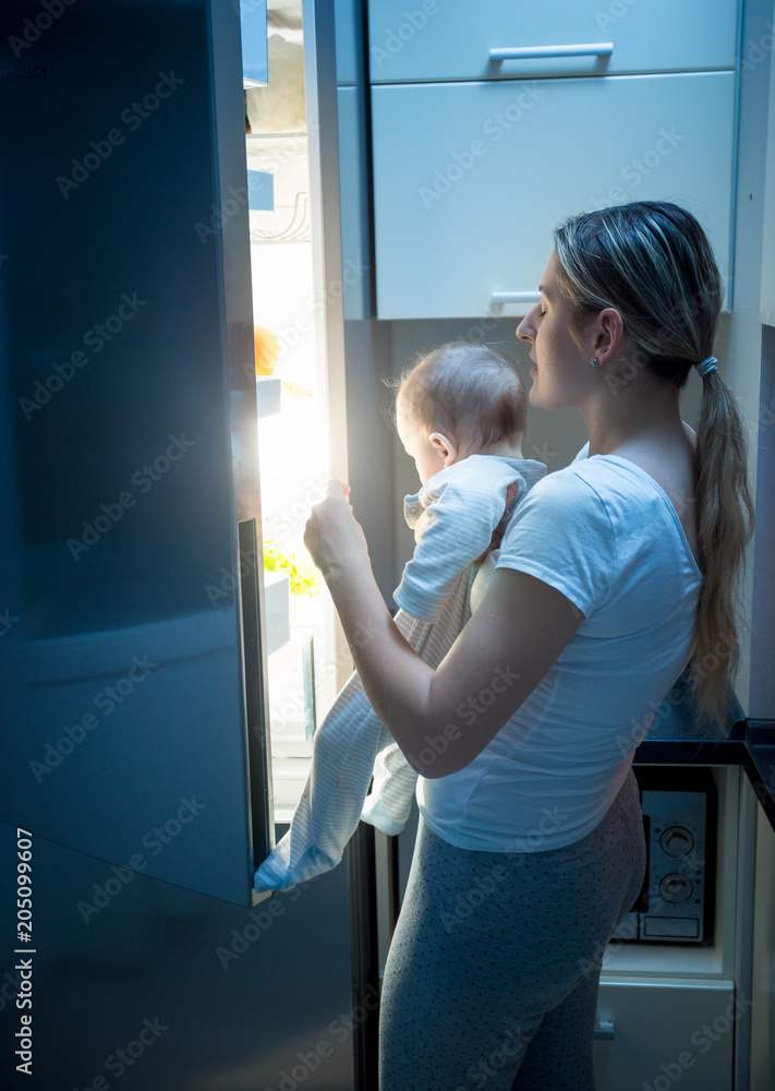 Portrait of young mother holding her baby opens refrigerator to find some food at night
