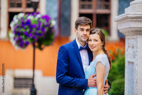 Beautiful newlyweds posing in old town. Adorable bride and groom kiss each other behind old orange wall under green tree leaves. Wedding day. Woman in blue dress. Man in suit. Young wedding couple. © nataliakabliuk
