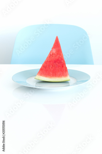 A slice of watermelon on a plate on a white table. A blue chair in the background.