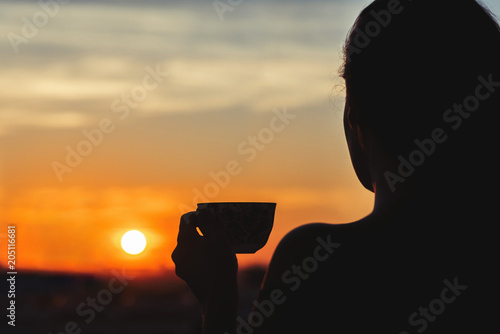  Silhouette of girl with a cup of tea at sunset. View on the town from above