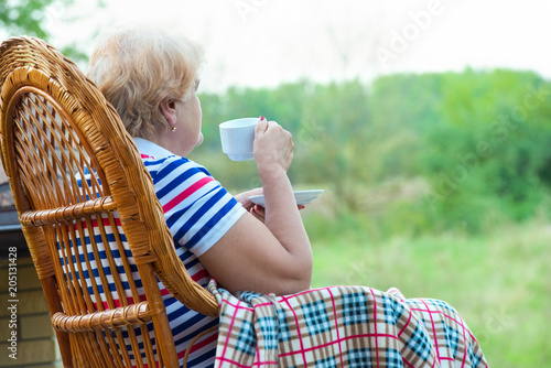 An elderly woman sits in a wicker rocking chair and and drinks a cup of hot tea.Relax in a country house.