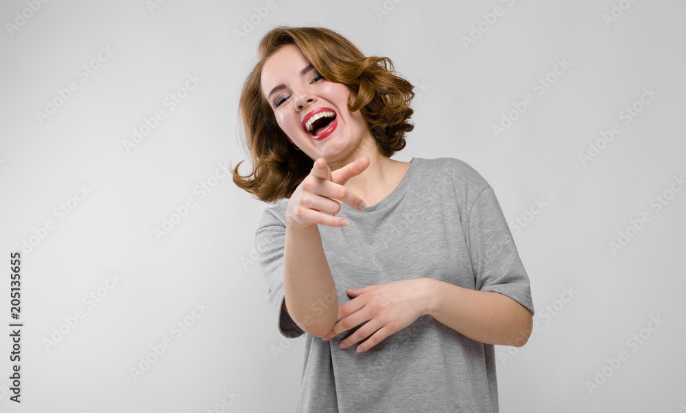 Charming young girl in a gray T-shirt on a gray background. The girl points with her finger and laughs Charming young girl in a gray T-shirt on a gray background. The girl points with her finger and laughs