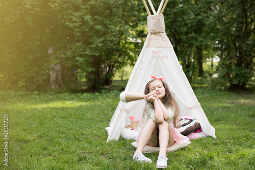 Little girl playing in a tent, children's house wigwam in park. Happy mothers day. Child with big donuts and ice cream and straw hat Summer, happy childhood concept. Cute emotions girl.