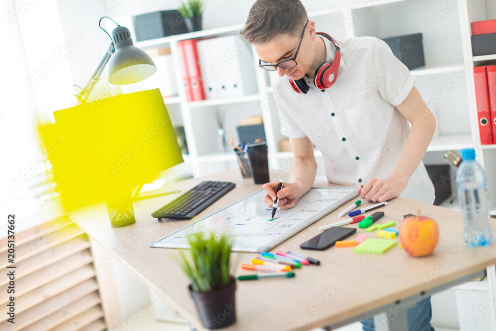 A young man in glasses stands near a computer desk. A young man draws a marker on a magnetic board. On the neck, the guy's headphones hang. A young man in glasses stands near a computer desk. A young man draws a marker on a magnetic board. On the neck, the guy's headphones hang.
