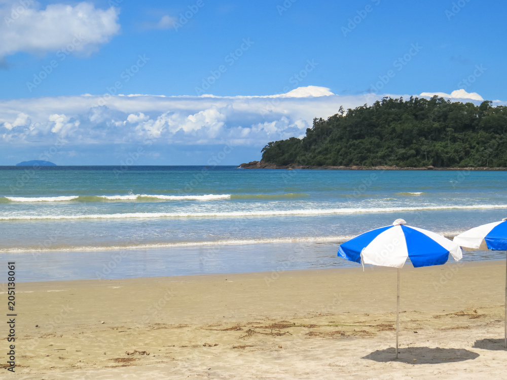 View of beach on sunny day with umbrella and island and clouds in the background.