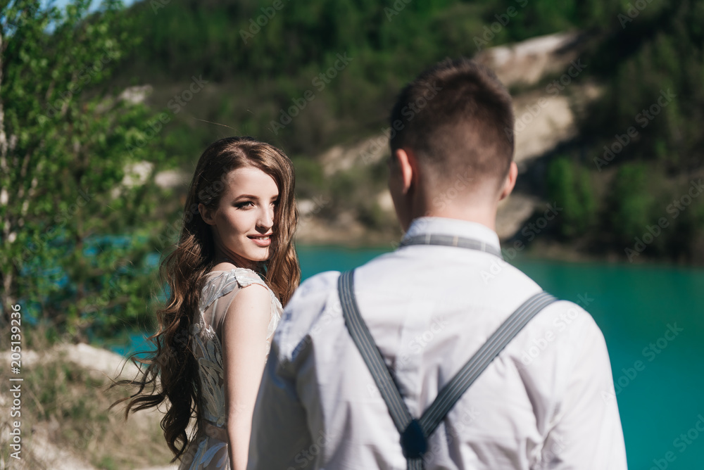 The bride in a beautiful dress holding hands with the groom in a light suit against the blue sky and blue water. Wedding couple standing on a sandy hill in the open air. A romantic love story.