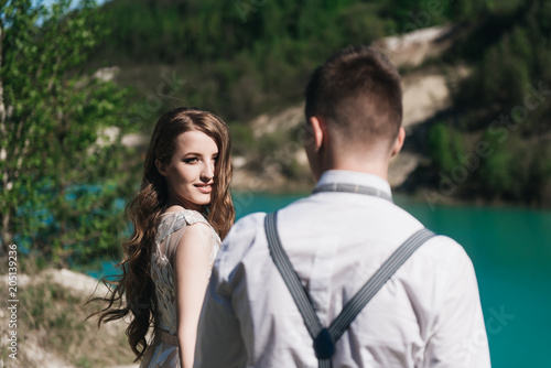 The bride in a beautiful dress holding hands with the groom in a light suit against the blue sky and blue water. Wedding couple standing on a sandy hill in the open air. A romantic love story.