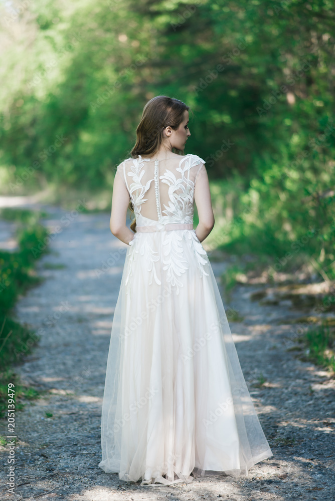 Beautiful brunette bride with curls, makeup and stylish dress. Portrait on the background of green trees. Summer wedding day.