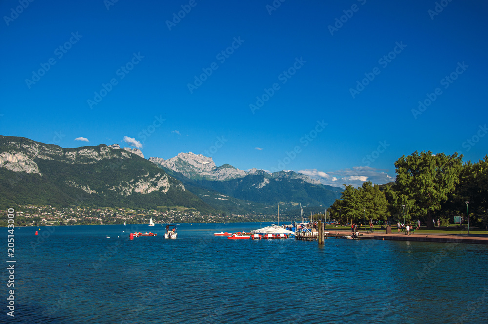 View of Annecy lake, with island, vegetation, paddle boats, mountains and blue sky in background. Located in the department of Haute-Savoie, Auvergne-Rhone-Alpes region, southeastern France.