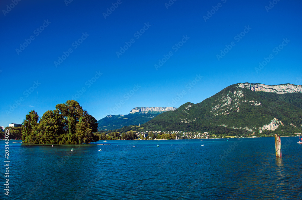View of Annecy lake, with island, vegetation, mountains and blue sky in background. Located in the department of Haute-Savoie, Auvergne-Rhone-Alpes region, southeastern France.