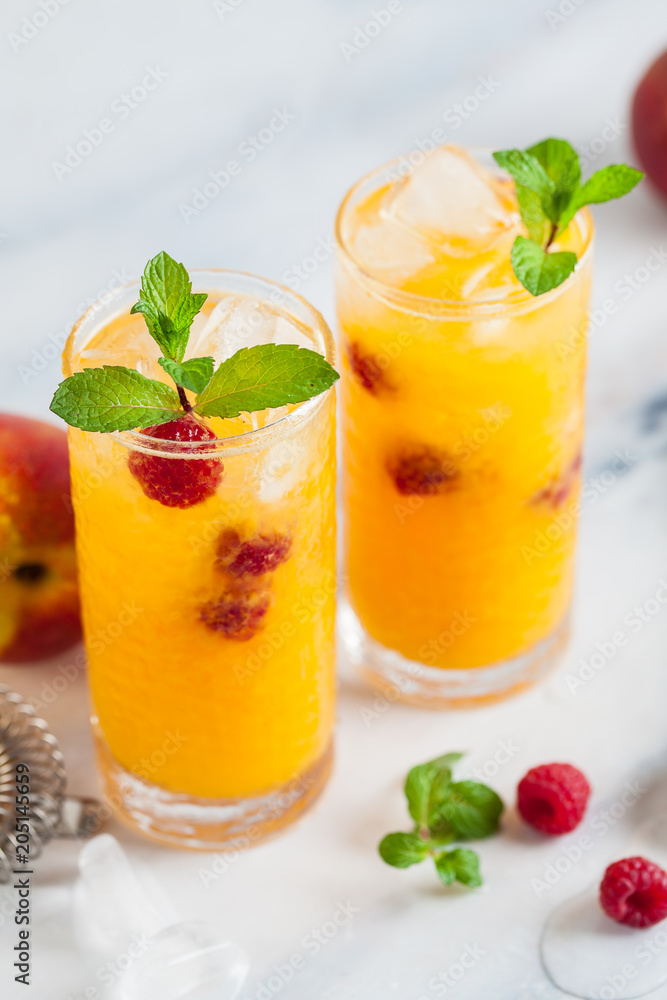 two refreshing drinks from peach juice and raspberries with fresh mint in tall glasses on a marble table with a shaker and bar accessories.