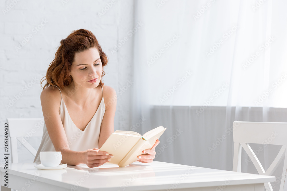 focused young woman reading book with coffee cup on table at home focused young woman reading book with coffee cup on table at home