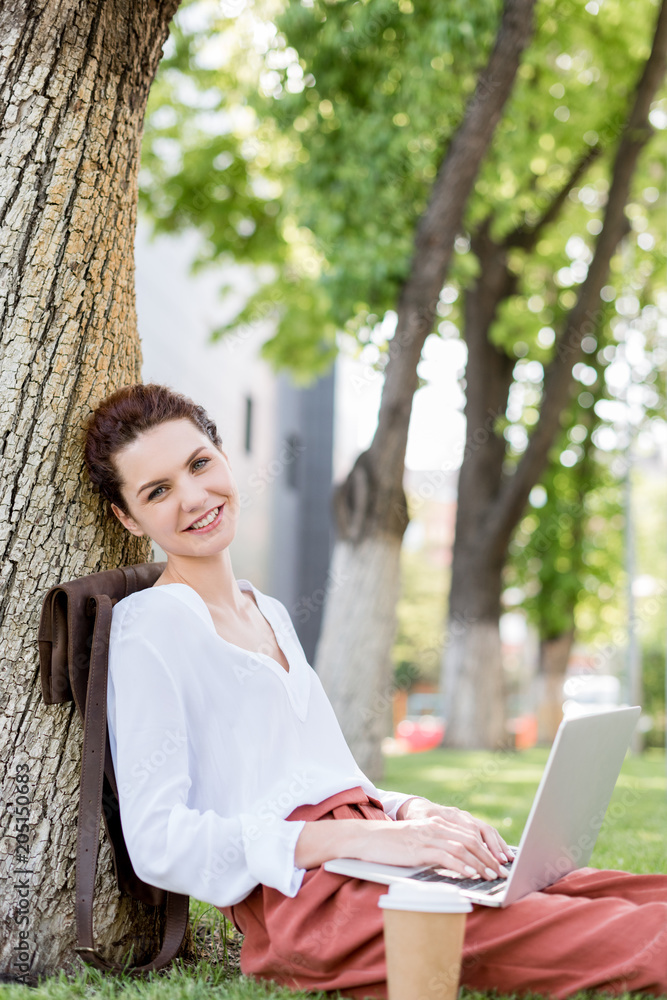 happy young freelancer working with laptop while leaning back on tree trunk in park and looking at camera happy young freelancer working with laptop while leaning back on tree trunk in park and looking at camera