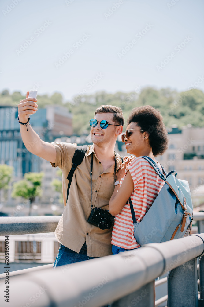 happy multicultural couple of tourists in sunglasses taking selfie on smartphone