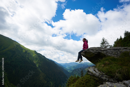 Silhouette of young female tourist sitting on the rock  dangling her legs in the abyss against the backdrop of green mountains with forests and clouds above them through which the blue sky is visible.
