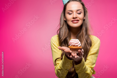 smiling attractive woman with party hat holding cupcake isolated on pink