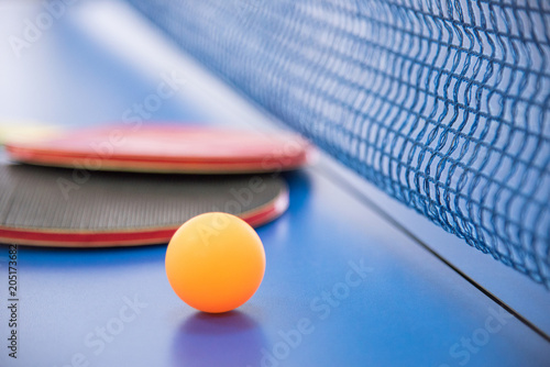 Orange ball for table tennis and two rackets of red and black color on a blue table with a grid