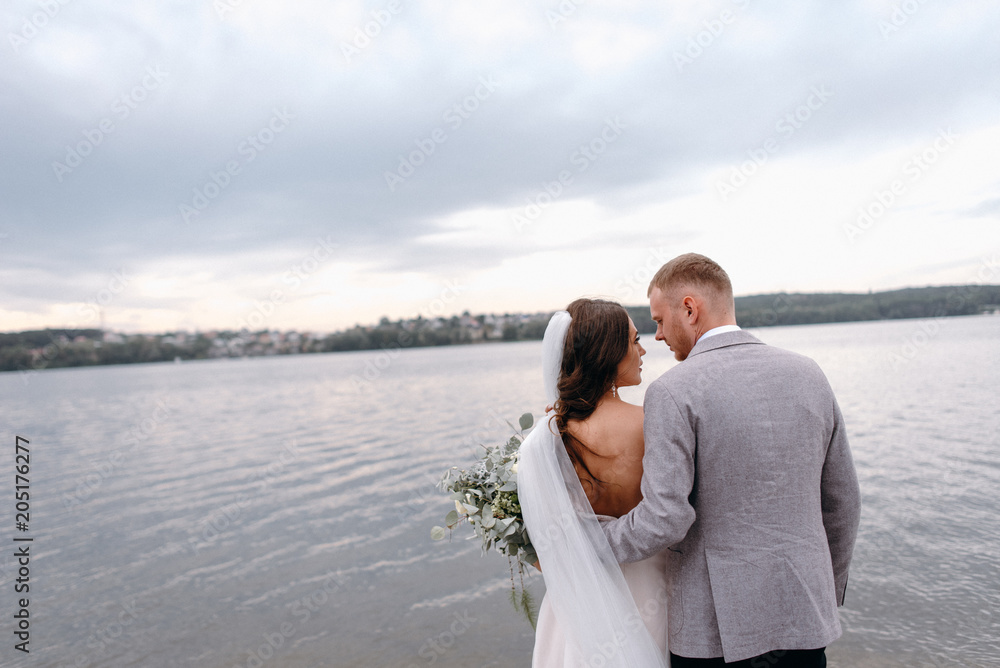 An amazing couple on their wedding day near the lake hug and enjoy each other. The bride and groom with a bouquet are happy together