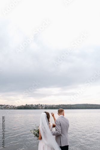 An amazing couple on their wedding day near the lake hug and enjoy each other. The bride and groom with a bouquet are happy together © Oleksandr