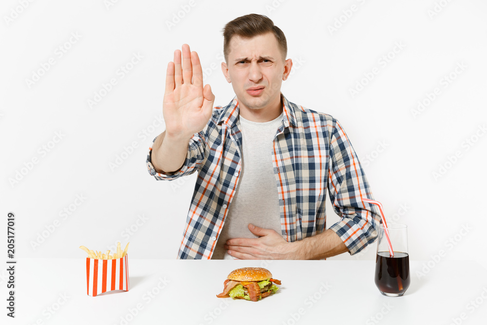 Illness man put hands on pain abdomen, stomach-ache at table with burger, french fries, cola in glass isolated on white background. Proper nutrition or American classic fast food. Area with copy space Illness man put hands on pain abdomen, stomach-ache at table with burger, french fries, cola in glass isolated on white background. Proper nutrition or American classic fast food. Area with copy space