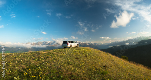 car stands on a mountain meadow with flowers and grass against the backdrop of the top of the mountain ranges snow-capped in the clouds